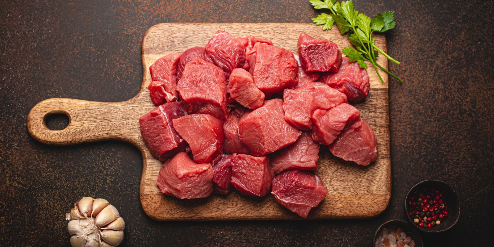 Chunks of red meat fill a wood cutting board, surrounded by sprigs of herbs, cups of spices and a whole garlic 